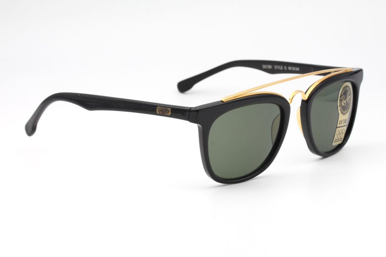 May include: Black sunglasses with a gold-colored metal bridge and dark green lenses. The sunglasses have a classic design with a rectangular frame and a curved temple. The text "Gatsby Style & Woman" is visible on the frame.