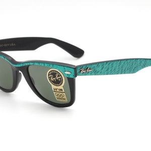 Puede incluir: Gafas de sol Ray-Ban Wayfarer negras con monturas verdes texturizadas. Las gafas de sol tienen lentes verdes y un logotipo Ray-Ban dorado en la patilla derecha. Las gafas de sol tienen una pegatina en la lente derecha que dice "G-15 Lens" y "Ray-Ban"