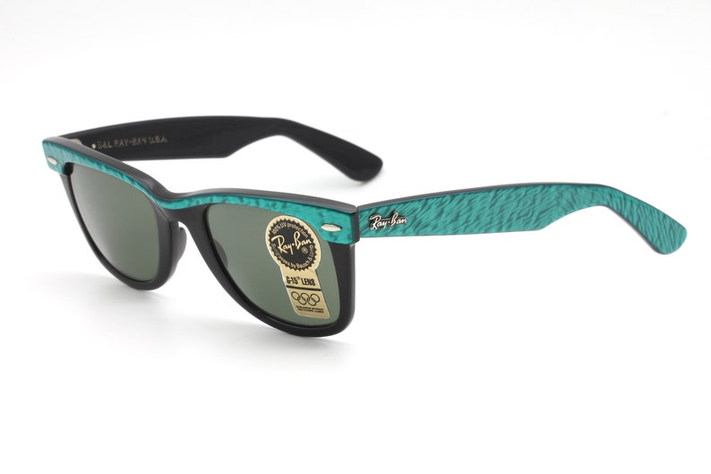 Puede incluir: Gafas de sol Ray-Ban Wayfarer negras con monturas verdes texturizadas. Las gafas de sol tienen lentes verdes y un logotipo Ray-Ban dorado en la patilla derecha. Las gafas de sol tienen una pegatina en la lente derecha que dice "G-15 Lens" y "Ray-Ban"