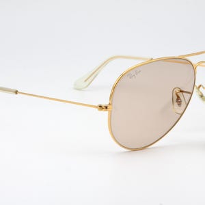 May include: Gold aviator sunglasses with brown tinted lenses and a Ray-Ban logo on the frame. The sunglasses are accompanied by a small, round, gold tag with the text "Original Bausch & Lomb" on a black background.