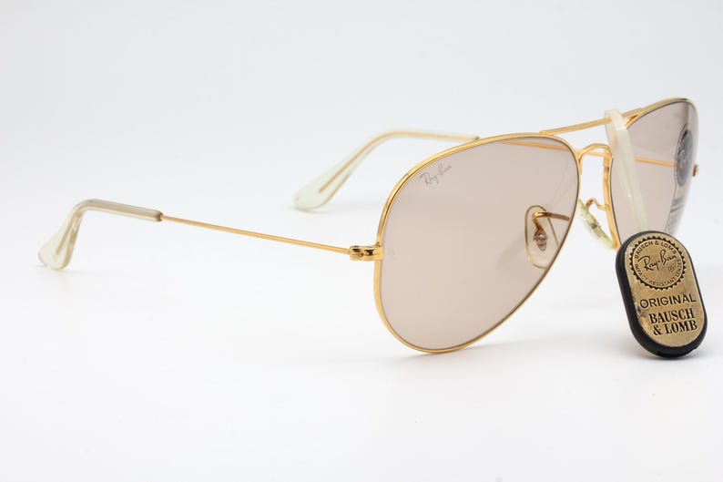 May include: Gold aviator sunglasses with brown tinted lenses and a Ray-Ban logo on the frame. The sunglasses are accompanied by a small, round, gold tag with the text "Original Bausch & Lomb" on a black background.