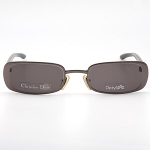 May include: Brown rectangular sunglasses with dark lenses. The sunglasses have a thin, metallic frame and gray temples. The brand name "Christian Dior" is visible on the left lens, and "Optyl" is on the right lens.