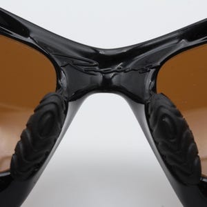 May include: Close-up of the bridge of a pair of black sunglasses with brown lenses. The sunglasses have a textured rubber detail on the arms.