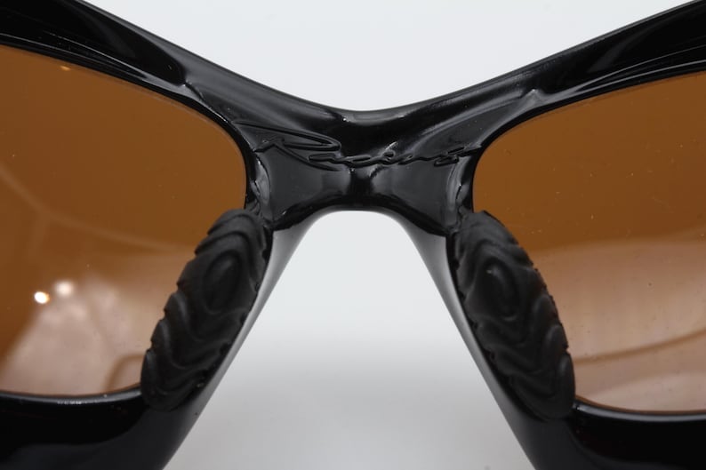 May include: Close-up of the bridge of a pair of black sunglasses with brown lenses. The sunglasses have a textured rubber detail on the arms.