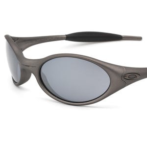 May include: Gray oval-shaped sunglasses with gray lenses. The sunglasses have a matte finish and a black arm. The Oakley logo is visible on the frame.