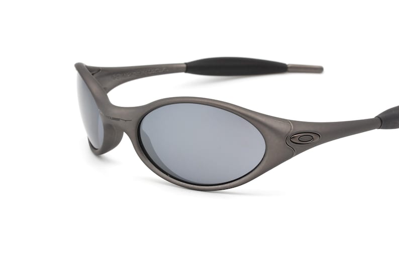 May include: Gray oval-shaped sunglasses with gray lenses. The sunglasses have a matte finish and a black arm. The Oakley logo is visible on the frame.