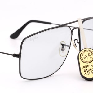 May include: Black Ray-Ban aviator sunglasses with a clear lens and a black metal frame. The sunglasses are sitting on a white surface. The sunglasses have a sticker on the side that says "Original Bausch & Lomb".