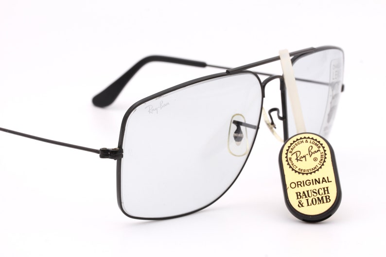 May include: Black Ray-Ban aviator sunglasses with a clear lens and a black metal frame. The sunglasses are sitting on a white surface. The sunglasses have a sticker on the side that says "Original Bausch & Lomb".