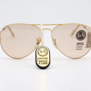 May include: Gold-framed aviator sunglasses with brown lenses. The sunglasses have a Ray-Ban logo on the right lens and a "CHANGEABLE" label with Olympic rings on the left lens. The sunglasses are sitting on a white surface.