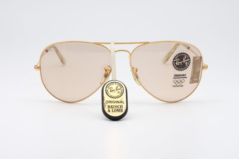 May include: Gold-framed aviator sunglasses with brown lenses. The sunglasses have a Ray-Ban logo on the right lens and a "CHANGEABLE" label with Olympic rings on the left lens. The sunglasses are sitting on a white surface.
