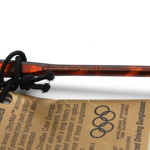 May include: Close-up of a brown and black tortoiseshell sunglasses frame with the text "BAL RAY BAN USA" printed on the side. The sunglasses are part of a set that includes a tag with the Olympic rings and the text "WORLDWIDE SPONSOR 1996 OLYMPIC GAMES" printed on it.
