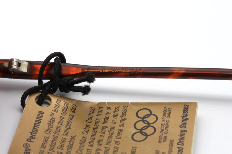 May include: Close-up of a brown and black tortoiseshell sunglasses frame with the text "BAL RAY BAN USA" printed on the side. The sunglasses are part of a set that includes a tag with the Olympic rings and the text "WORLDWIDE SPONSOR 1996 OLYMPIC GAMES" printed on it.