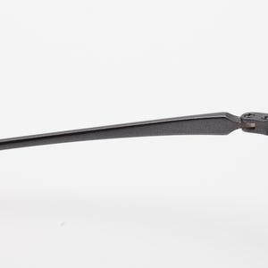 May include: Close-up of a pair of eyeglasses with a dark gray, matte finish. The frame has a thin, rectangular shape with a hinge connecting the temple arm to the front of the frame. The lenses are clear.