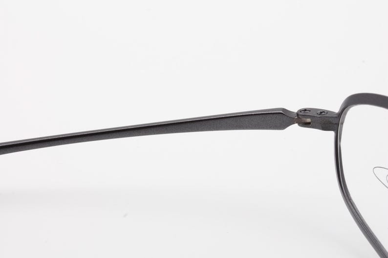 May include: Close-up of a pair of eyeglasses with a dark gray, matte finish. The frame has a thin, rectangular shape with a hinge connecting the temple arm to the front of the frame. The lenses are clear.