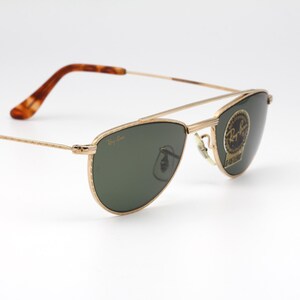 May include: Gold-framed sunglasses with green lenses. The sunglasses have a double bridge and a classic aviator shape. The temples are a tortoiseshell pattern. The Ray-Ban logo is visible on the lens.