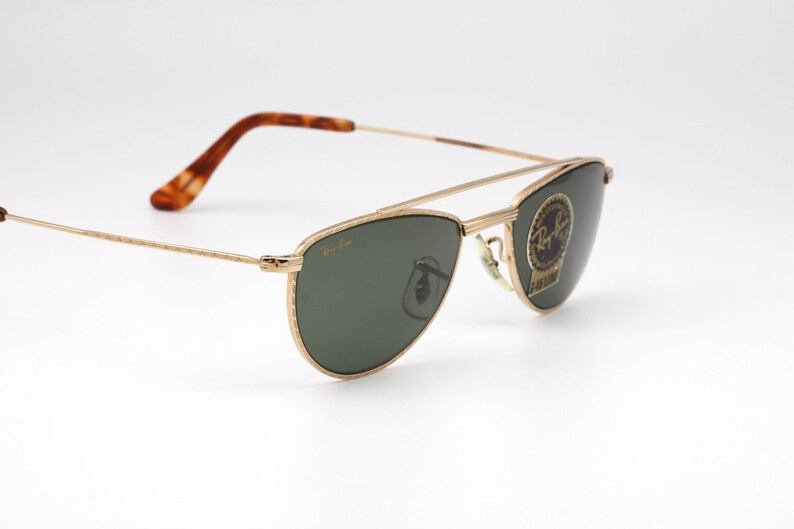 May include: Gold-framed sunglasses with green lenses. The sunglasses have a double bridge and a classic aviator shape. The temples are a tortoiseshell pattern. The Ray-Ban logo is visible on the lens.