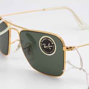 May include: Gold-tone Ray-Ban sunglasses with green lenses. The sunglasses have a square frame and a white nose bridge. The Ray-Ban logo is visible on the right lens.