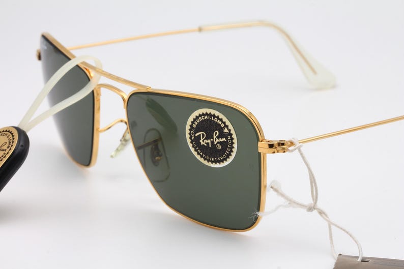 May include: Gold-tone Ray-Ban sunglasses with green lenses. The sunglasses have a square frame and a white nose bridge. The Ray-Ban logo is visible on the right lens.