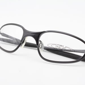 May include: Black eyeglasses with a sleek, modern design. The frames are a matte black, with clear lenses. The Oakley logo is visible on the lens. The glasses are set against a white background.
