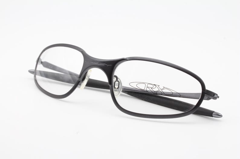 May include: Black eyeglasses with a sleek, modern design. The frames are a matte black, with clear lenses. The Oakley logo is visible on the lens. The glasses are set against a white background.