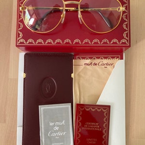 May include: A gold-rimmed pair of glasses with a red leather case and a brown leather case with the words 'les must de Cartier Paris' and 'CERTIFICAT DE GARANTIE INTERNATIONALE LUNETTES CARTIER' printed on the cases.