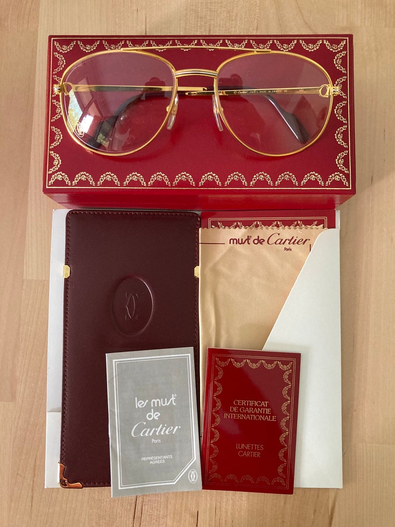 May include: A gold-rimmed pair of glasses with a red leather case and a brown leather case with the words 'les must de Cartier Paris' and 'CERTIFICAT DE GARANTIE INTERNATIONALE LUNETTES CARTIER' printed on the cases.