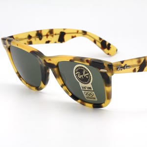 Puede incluir: Gafas de sol con estampado de carey y lentes oscuras. Las monturas son amarillas y marrones, con el logo de Ray-Ban en la patilla. Una pegatina con el logo y texto de Ray-Ban est&aacute; en la lente.