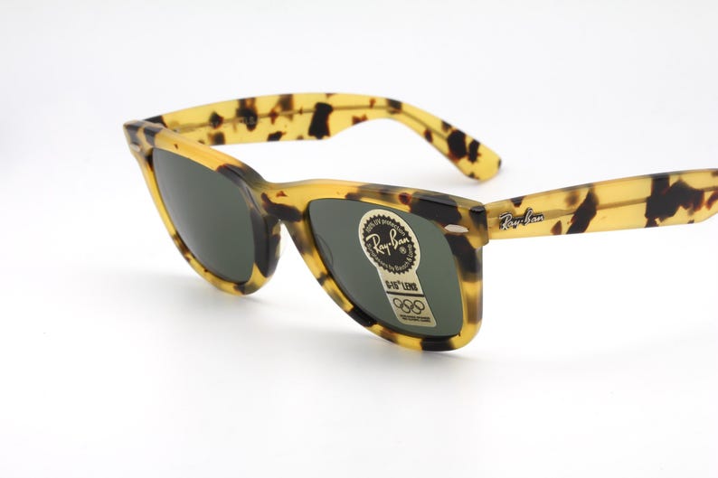 Puede incluir: Gafas de sol con estampado de carey y lentes oscuras. Las monturas son amarillas y marrones, con el logo de Ray-Ban en la patilla. Una pegatina con el logo y texto de Ray-Ban est&aacute; en la lente.