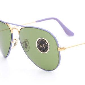 Op de afbeelding: Paarse en gouden aviator zonnebril met groene glazen. De zonnebril heeft een Ray-Ban logo sticker op de rechter lens.