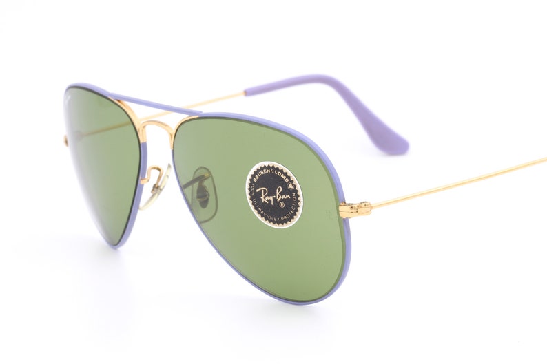 Op de afbeelding: Paarse en gouden aviator zonnebril met groene glazen. De zonnebril heeft een Ray-Ban logo sticker op de rechter lens.