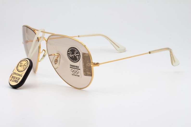 May include: Gold-framed aviator sunglasses with brown lenses. The sunglasses feature a Ray-Ban logo and the text "CHANGEABLE" on the right lens. The sunglasses are accompanied by a small, round sticker with the text "ORIGINAL BAUSCH & LOMB".