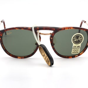 Op de afbeelding: Schildpad Ray-Ban zonnebril met groene glazen en een gouden metalen frame. De zonnebril heeft een wit koord dat aan het frame is bevestigd en een gouden sticker met de tekst "Ray-Ban G-15 Lens" en de Olympische ringen.