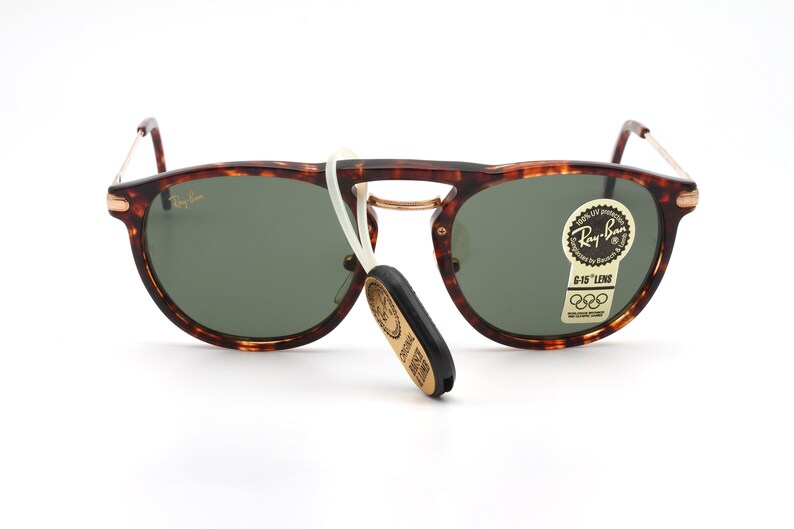 Op de afbeelding: Schildpad Ray-Ban zonnebril met groene glazen en een gouden metalen frame. De zonnebril heeft een wit koord dat aan het frame is bevestigd en een gouden sticker met de tekst "Ray-Ban G-15 Lens" en de Olympische ringen.