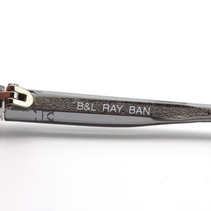 May include: Close-up of the arm of a pair of gray sunglasses with a silver hinge and the text "B&L RAY BAN" printed on the arm.