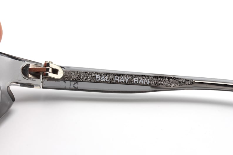 May include: Close-up of the arm of a pair of gray sunglasses with a silver hinge and the text "B&L RAY BAN" printed on the arm.