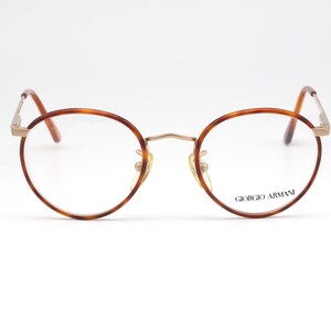 May include: Round eyeglasses with a brown tortoiseshell frame and gold-colored metal accents. The lenses are clear, and the brand name "GIORGIO ARMANI" is printed on the right lens. The glasses have a classic, vintage style.