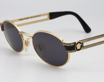 Puede incluir: Gafas de sol ovaladas doradas con lentes negras y un diseño de llave griega en negro y dorado en las patillas. Las gafas de sol tienen el logotipo de Versace en la patilla derecha.
