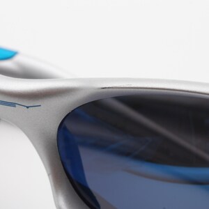 May include: Silver and blue Oakley sunglasses with a blue frame and blue lenses. The Oakley logo is visible on the frame.