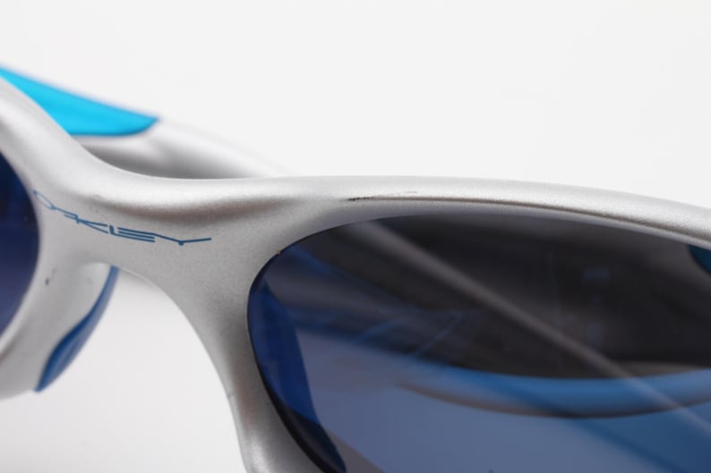 May include: Silver and blue Oakley sunglasses with a blue frame and blue lenses. The Oakley logo is visible on the frame.