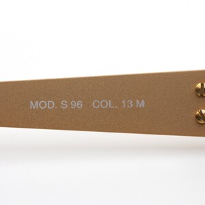 May include: Close-up of the gold-toned metal arm of a pair of sunglasses. The arm is engraved with the text "MOD. S 96 COL. 13 M".