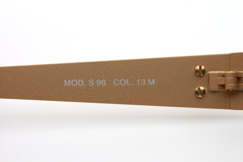 May include: Close-up of the gold-toned metal arm of a pair of sunglasses. The arm is engraved with the text "MOD. S 96 COL. 13 M".