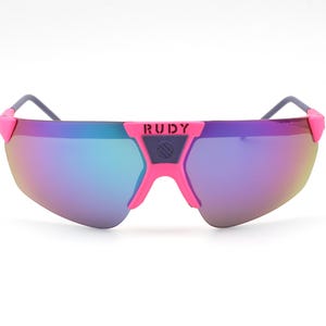 May include: Pink and purple sunglasses with a mirrored lens. The sunglasses have a pink frame with the brand name "RUDY" in white letters. The lenses have a gradient of colours, including blue, purple, and yellow.