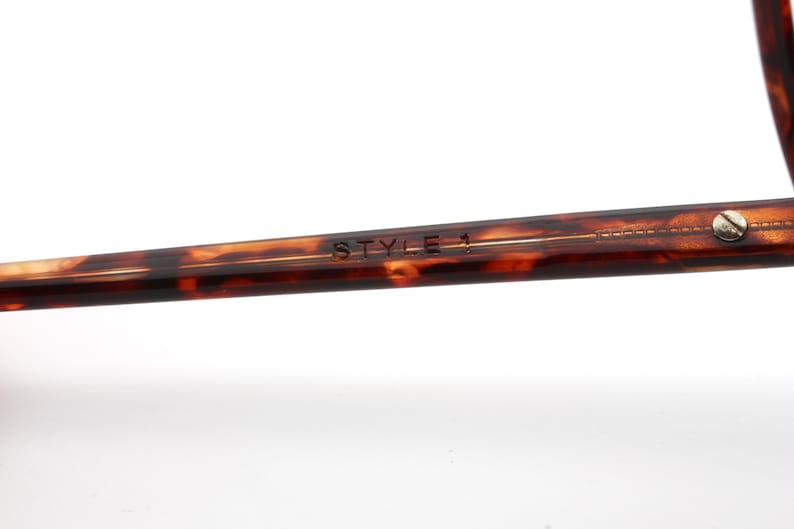 May include: Close-up of the arm of eyeglasses with a tortoiseshell pattern. The text "STYLE 1" is engraved on the arm. A small silver screw is visible near the end of the arm. The glasses are set against a white background.