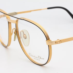 May include: Gold-tone eyeglasses with a black top bar and temples. The frames have a classic aviator shape with a black outline. The lenses are clear, and the brand name "Christian Dior" is visible on the lens.