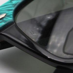 Puede incluir: Primer plano de unas gafas de sol negras y verdes con una montura verde texturizada. Las gafas de sol tienen una montura de pl&aacute;stico negro y lentes verdes. El texto "22" es visible en la lente derecha.