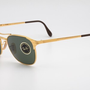 May include: Gold-tone Ray-Ban sunglasses with green lenses. The sunglasses have a classic aviator style with a thin metal frame and brown plastic temples.