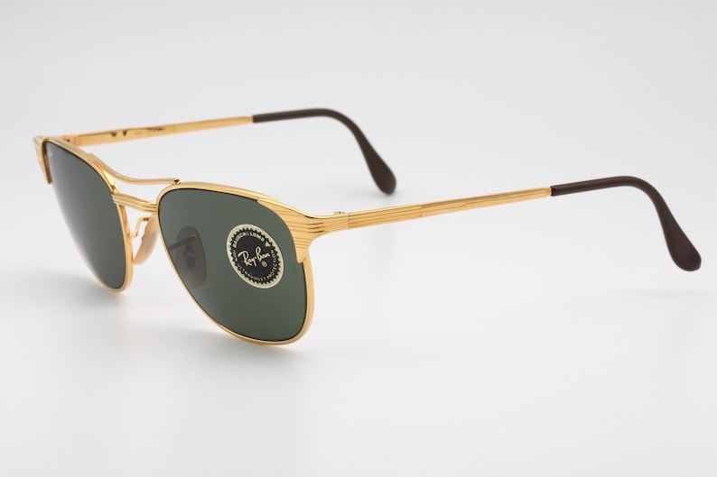 May include: Gold-tone Ray-Ban sunglasses with green lenses. The sunglasses have a classic aviator style with a thin metal frame and brown plastic temples.
