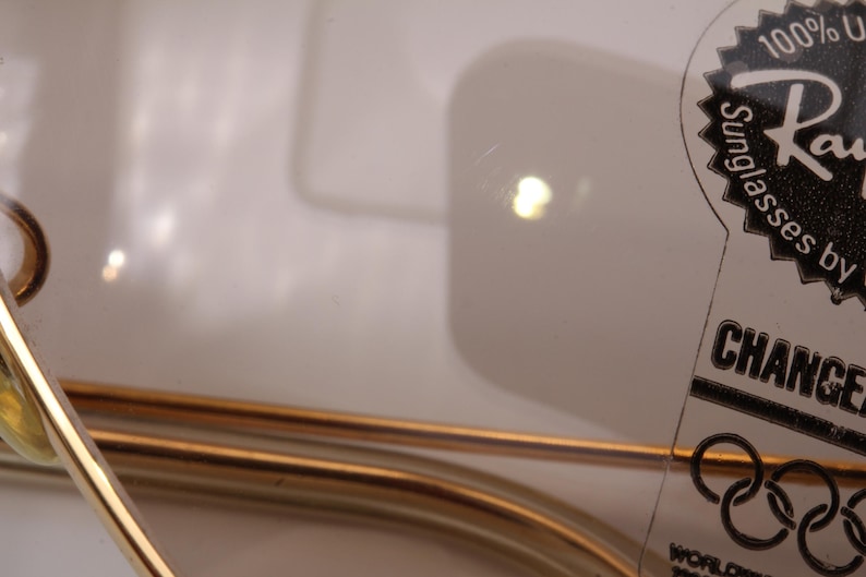 May include: Close-up of a gold-coloured metal frame of a pair of sunglasses with a black and white label that reads "100% UV Sunglasses by Ray-Ban CHANGE"