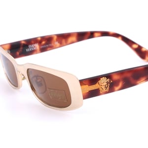 Puede incluir: Gafas de sol marrones y doradas con montura de carey y lentes marrones. Las gafas tienen el texto "GIANNI VERSACE" en la montura.
