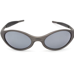 May include: Gray oval-shaped sunglasses with gray lenses. The sunglasses have a dark gray frame and temples. The brand name is subtly printed on the frame. A modern, sporty eyewear accessory.